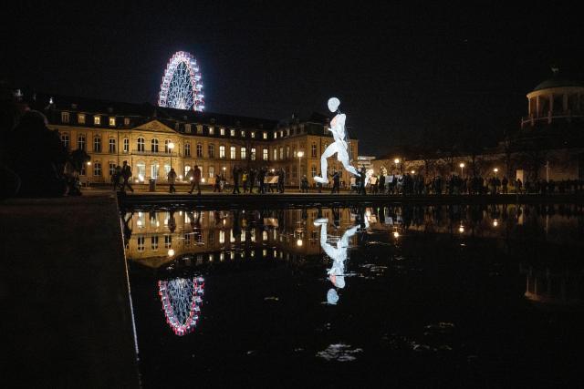01 December 2025, Baden-Württemberg, Stuttgart: The eight meter high illuminated puppet "Vektor" from the large puppet theater "Dundu" is led by puppeteers at the Eckensee in front of the New Palace in Stuttgart. The performance of the large puppet accompanied a campaign by AIDS-Hilfe Stuttgart to mark World AIDS Day. Photo: Marijan Murat/dpa