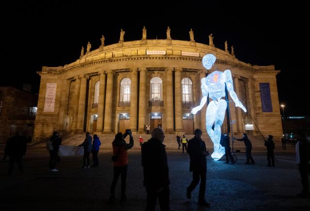 01 December 2025, Baden-Württemberg, Stuttgart: The eight meter high illuminated puppet "Vektor" from the large puppet theater "Dundu" is led by puppeteers in front of the State Opera in Stuttgart. The performance of the large puppet accompanied a campaign by AIDS-Hilfe Stuttgart to mark World AIDS Day. Photo: Marijan Murat/dpa