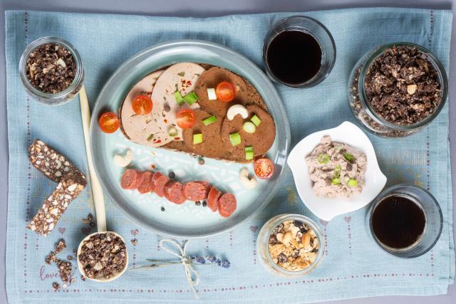 FILED - 06 January 2024, Saxony, Dresden: A slice of bread topped with various vegan sausages, a vegan garlic sausage and a bowl with a vegan pea-based liver sausage alternative, muesli with a plant-based yoghurt alternative, cashew date bars and coffee on a table. Photo: Sebastian Kahnert/dpa