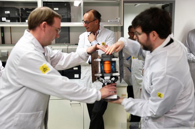 03 December 2025, Berlin: Engineers hand over a satellite to German Chancellor Friedrich Merz (C) during his visit to Berlin Space Technologies (BST) in Adlershof. Photo: Nadja Wohlleben/Reuters/Pool/dpa
