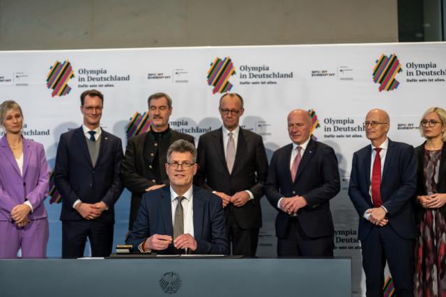 04 December 2025, Berlin: Thomas Weikert, President of the German Olympic Sports Confederation (DOSB) sits in front of (L-R) Britta Heidemann, IOC member, Hendrik Wuest, Minister President of North Rhine-Westphalia, Markus Soeder, Minister President of Bavaria and CSU Chairman, German Chancellor Friedrich Merz, Kai Wegner, Governing Mayor of Berlin, Peter Tschentscher, First Mayor of Hamburg, and Christiane Schenderlein, Minister of State for Sport and Volunteering, at the signing of the agreement on Germany's bid to host the Olympic Games in Germany. Photo: Michael Kappeler/dpa