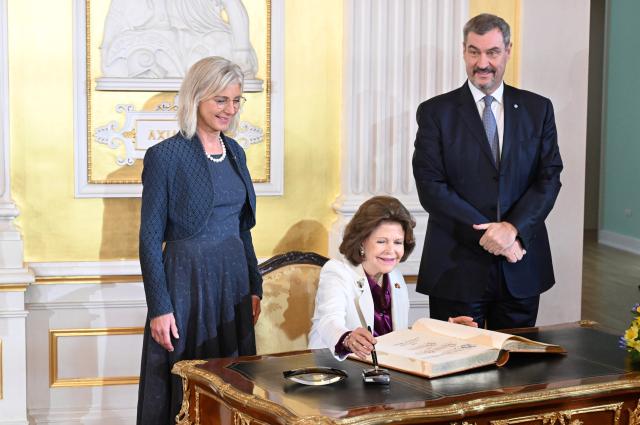 17 December 2025, Bavaria, Munich: Queen Silvia of Sweden signs the guest book of the state government in the Munich Residence, while Ulrike Scharf (L), Minister of State in the State Ministry for Family Affairs, Labor and Social Affairs and Bavarian Prime Minister Markus Soeder (R) look on. Photo: Felix Hörhager/dpa