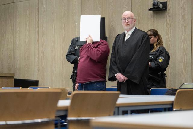 18 December 2025, Baden-Württemberg, Mannheim: The defendant (front left) in a murder trial stands with his lawyer Uwe Kosmala (2nd from right) and court officials in a courtroom at Mannheim Regional Court, covering his face. According to the public prosecutor's office, the man deliberately drove towards people during a drive at the beginning of March. The public prosecutor's office sees no evidence of a political motive. Photo: Marijan Murat/dpa