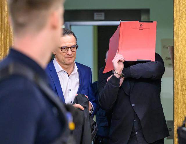 14 January 2026, Brandenburg, Cottbus: The defendant (R) arrives handcuffed at the Cottbus Regional Court, shielding his face with a file folder and accompanied by his defence attorney Christoph Ruehlmann, as the murder trial begins. The case concerns the death of a Dresden police officer who was fatally struck by a car during an operation against fleeing car thieves in southern Brandenburg. Photo: Patrick Pleul/dpa