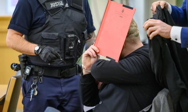 14 January 2026, Brandenburg, Cottbus: The defendant (C) shields his face with a file folder at the Cottbus Regional Court as the murder trial begins. The case concerns the death of a Dresden police officer who was fatally struck by a car during an operation against fleeing car thieves in southern Brandenburg. Photo: Patrick Pleul/dpa