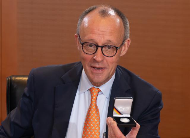 14 January 2026, Berlin: Germany's Chancellor Friedrich Merz shows a commemorative coin at the Chancellery at the start of the cabinet meeting. The two-euro coin is part of the "Unity, Justice, and Freedom" series. It was issued on the occasion of Konrad Adenauer's 150th birthday in a limited edition of 30 million and distributed to all members of the cabinet at the cabinet meeting. Photo: Soeren Stache/dpa