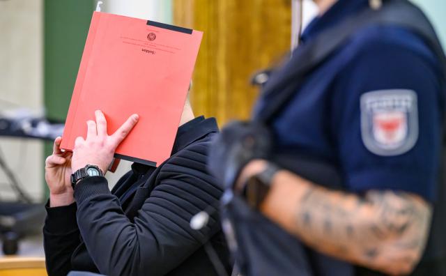 14 January 2026, Brandenburg, Cottbus: The defendant (L) shields his face with a file folder at the Cottbus Regional Court, as the murder trial begins. The case concerns the death of a Dresden police officer who was fatally struck by a car during an operation against fleeing car thieves in southern Brandenburg. Photo: Patrick Pleul/dpa