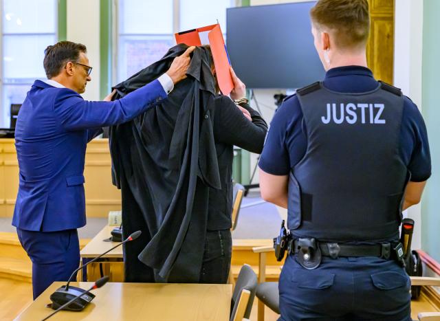 14 January 2026, Brandenburg, Cottbus: The defendant (C) arrives handcuffed at the Cottbus Regional Court, shielding his face with a file folder and accompanied by his defence attorney Christoph Ruehlmann, as the murder trial begins. The case concerns the death of a Dresden police officer who was fatally struck by a car during an operation against fleeing car thieves in southern Brandenburg. Photo: Patrick Pleul/dpa
