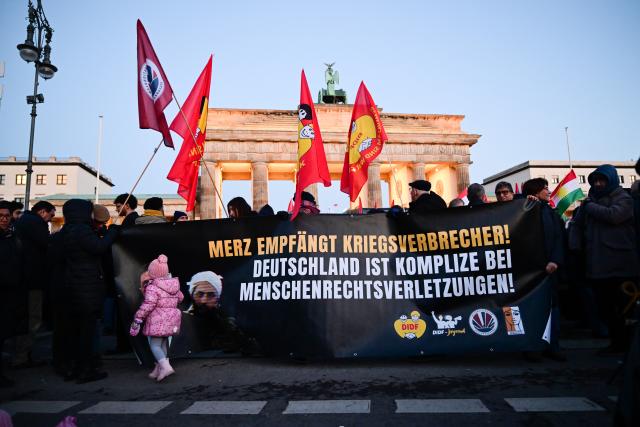 19 January 2026, Berlin: People hold a banner says "Merz welcomes war criminals! Germany is complicit in human rights violations!" during a demonstration with the slogan "No welcome for Islamist Ahmed al-Sharaa in Berlin" against the visit of the Syrian interim president. The two-day visit planned for today has been postponed. Photo: Sebastian Christoph Gollnow/dpa