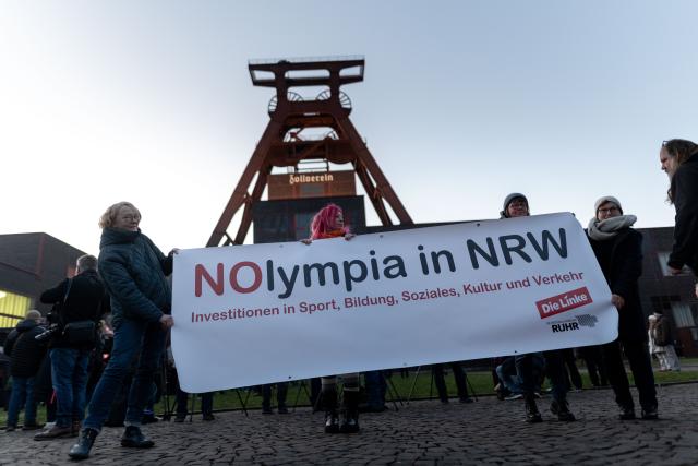 19 January 2026, North Rhine-Westphalia, Essen: People protest against the Olympic Games being held in North Rhine-Westphalia on the grounds of the Zollverein Coal Mine Industrial Complex, holding up a banner reading "NOlympia in NRW." A light installation by Bielefeld artist Hans-Christoph Muecke is projected onto the Zollverein Coal Mine's double trestle in Essen to mark the start of the campaign for North Rhine-Westphalia's Olympic bid. Photo: Fabian Strauch/dpa