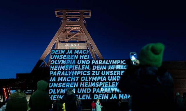 19 January 2026, North Rhine-Westphalia, Essen: A man holds a cell phone in the air and takes a photo at the Zollverein Coal Mine Industrial Complex in Essen, where a light installation by Bielefeld artist Hans-Christoph Muecke is projected onto the double trestle to mark the start of the campaign for an Olympic bid in North Rhine-Westphalia. Photo: Fabian Strauch/dpa
