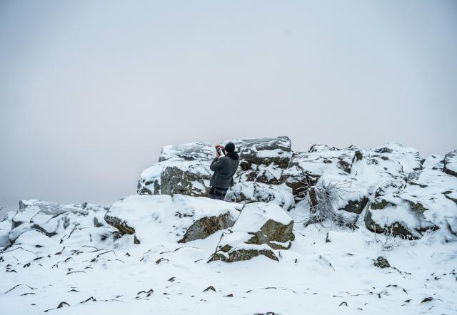 27 January 2026, Hesse, Schmitten: A man stands on the Brunhildisfelsen and takes a photo of the wintry landscape. The Feldberg plateau is covered in clouds with light snow or sleet and temperatures just below zero degrees Celsius. Photo: Andreas Arnold/dpa