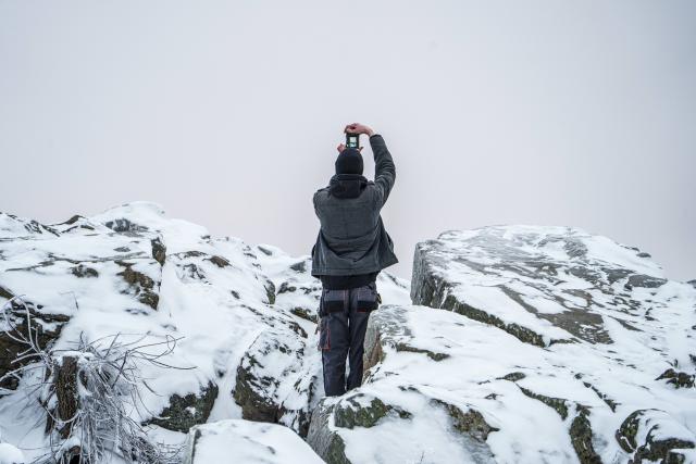 27 January 2026, Hesse, Schmitten: A man stands on the Brunhildisfelsen and takes a photo of the wintry landscape. The Feldberg plateau is covered in clouds with light snow or sleet and temperatures just below 0°. Photo: Andreas Arnold/dpa