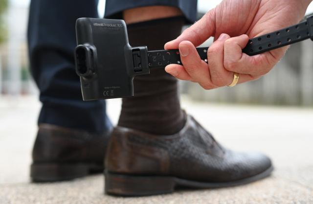 FILED - 04 August 2023, Hesse, Weiterstadt: A man holds an electronic monitoring device, known as an electronic ankle bracelet, in front of a test subject's leg for demonstration purposes. Photo: Arne Dedert/dpa