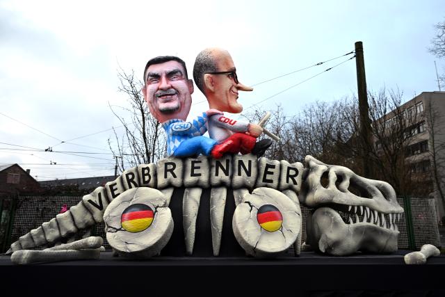 16 February 2026, North Rhine-Westphalia, Cologne: A themed float with Merz and Soeder riding the dinosaur Verbrenner drives out of the float construction hall. Rose Monday on the Rhine is the highlight of the Rhineland carnival season with its traditional parades. Photo: Federico Gambarini/dpa