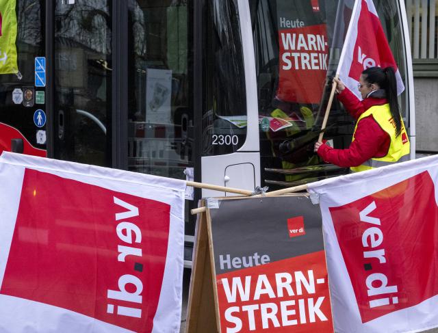 19 February 2026, Bavaria, Augsburg: A woman hangs a Verdi union flag next to a warning strike notice. Today, there will be considerable restrictions and cancellations in local public transport as part of the all-day warning strikes. Photo: Stefan Puchner/dpa