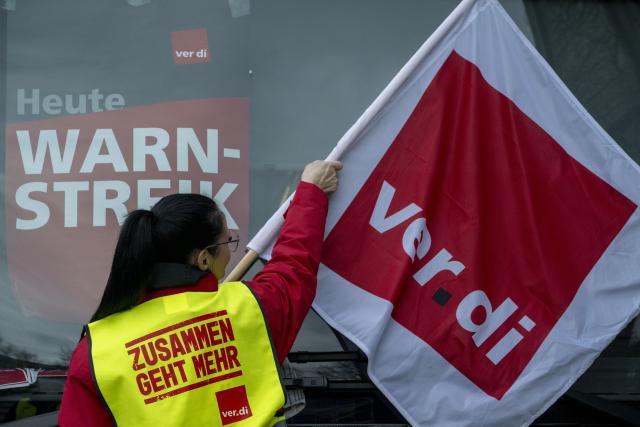 19 February 2026, Bavaria, Augsburg: A woman hangs a Verdi union flag next to a warning strike notice. Today, there will be considerable restrictions and cancellations in local public transport as part of the all-day warning strikes. Photo: Stefan Puchner/dpa
