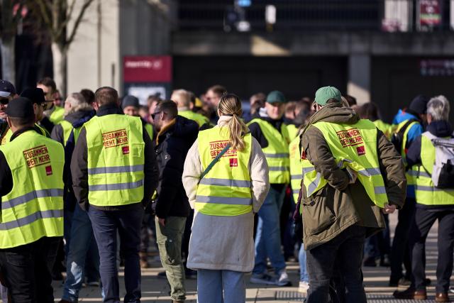 18 March 2026, Brandenburg, Schoenefeld: Striking airport employees and Verdi union members stand in front of the terminal during a warning strike at Berlin Brandenburg Airport (BER), halting all operations. No passenger flights have taken off or landed at BER since this morning, with all departures and arrivals cancelled for the entire day, the Berlin-Brandenburg Airport Company (FBB) had announced. Photo: Michael Ukas/dpa