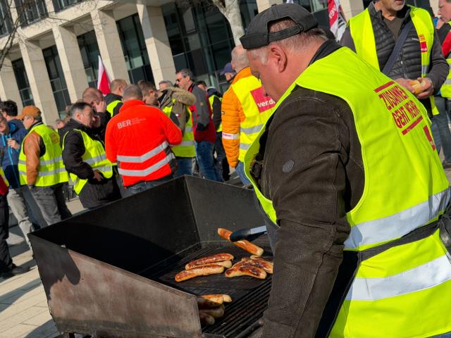 18 March 2026, Brandenburg, Schoenefeld: Striking airport employees and Verdi union members stand in front of the terminal during a warning strike at Berlin Brandenburg Airport (BER), halting all operations. No passenger flights have taken off or landed at BER since this morning, with all departures and arrivals cancelled for the entire day, the Berlin-Brandenburg Airport Company (FBB) had announced. Photo: Julian Degler/dpa