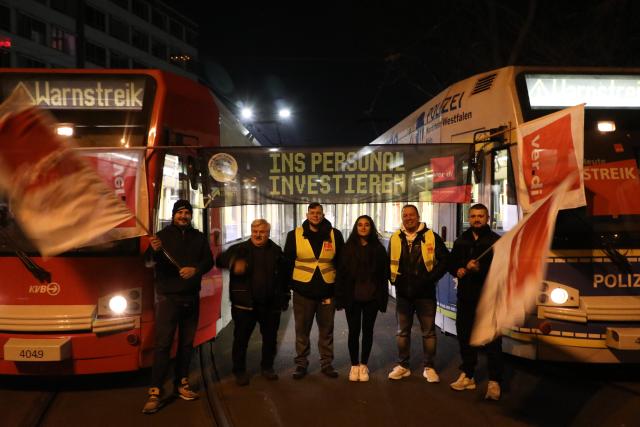 19 March 2026, North Rhine-Westphalia, Cologne: Strikers stand with flags in front of parked trams during a warning strike. As part of ongoing collective bargaining negotiations for local public transportation, the Verdi union has called for warning strikes to take place simultaneously in several states on Thursday. Photo: Sascha Thelen/dpa