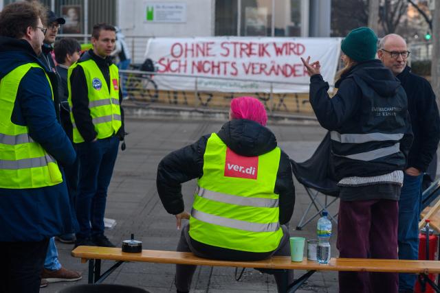 19 March 2026, Saxony-Anhalt, Magdeburg: "Nothing will change without a strike" can be read on a fabric banner hanging on a railing behind strikers. The trade union Verdi has once again called for warning strikes in local public transport in several federal states. In Magdeburg, some streetcar and bus lines are running in emergency mode. Photo: Klaus-Dietmar Gabbert/dpa
