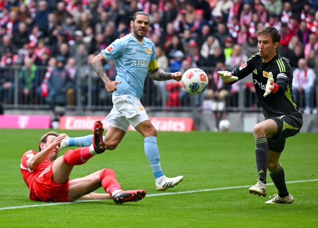 21 March 2026, Bavaria, Munich: Bayern Munich's Harry Kane (L) battle for the ball with Union Berlin's Christopher Trimmel (C) and goalkeeper Frederik Roennow during the German Bundesliga soccer match between Bayern Munich and 1. FC Union Berlin at the Allianz Arena. Photo: Sven Hoppe/dpa - WICHTIGER HINWEIS: Gemäß den Vorgaben der DFL Deutsche Fußball Liga bzw. des DFB Deutscher Fußball-Bund ist es untersagt, in dem Stadion und/oder vom Spiel angefertigte Fotoaufnahmen in Form von Sequenzbildern und/oder videoähnlichen Fotostrecken zu verwerten bzw. verwerten zu lassen.