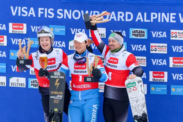 21 March 2026, North Rhine-Westphalia, Winterberg: (L-R) Austria's second placed Christoph Karner, South Korea's winner Lee Sang-ho and Austria's third placed Alexander Payer celebrate on the podium after the Men's Parallel Slalom of the FIS Snowboard World Cup in Winterberg. Photo: Christoph Reichwein/dpa