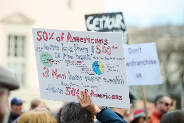 28 March 2026, Berlin: A protester holds a sign reading '50% of Americans have less than $500... Three men have more wealth than 50% of Americans' on Bebelplatz during a demonstration against the Trump administration. Protests against President Donald Trump's policies are taking place in Germany and in many US cities as well as other locations worldwide. Photo: Annette Riedl/dpa