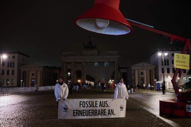28 March 2026, Berlin: A banner reading 'Off with fossil fuels. On with renewables.' hangs beneath an oversized lamp in front of the dark Brandenburg Gate during Earth Hour. The global campaign, organized by the environmental group WWF, sees lights switched off for one hour from 20:30 local time to raise awareness of climate and environmental protection. Photo: Annette Riedl/dpa