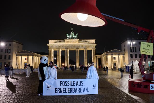 28 March 2026, Berlin: A banner reading 'Off with fossil fuels. On with renewables.' hangs beneath an oversized lamp in front of the dark Brandenburg Gate during Earth Hour. The global campaign, organized by the environmental group WWF, sees lights switched off for one hour from 20:30 local time to raise awareness of climate and environmental protection. Photo: Annette Riedl/dpa