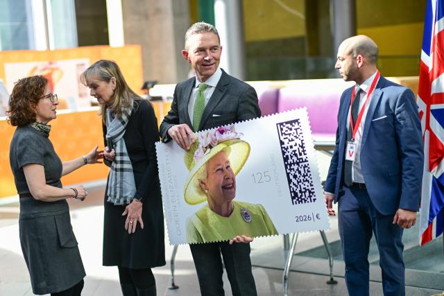 02 April 2026, Berlin: British Ambassador to the Federal Republic of Germany Andrew Mitchell (C) holds an oversized replica of the stamp in his arms during the unveiling of a commemorative stamp marking "the 100th birthday of Her Majesty Queen Elizabeth II" at the British Embassy. Photo: Sebastian Christoph Gollnow/dpa