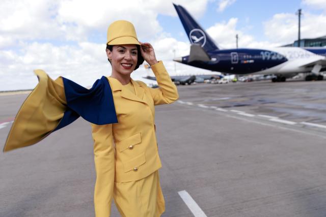 06 April 2026, Brandenburg, Schoenefeld: A hostess wears a former Lufthansa uniform in front of a Lufthansa passenger aircraft at Berlin Brandenburg Airport (BER) during celebrations marking the 100th anniversary of the first flight. Photo: Carsten Koall/dpa