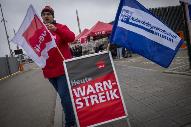 14 April 2026, Bavaria, Munich: Employees of the MVG East Depot stand behind a banner reading "Warning Strike Today" at the workshop gate on Truderinger Strasse during a warning strike affecting local public transportation. Photo: Peter Kneffel/dpa