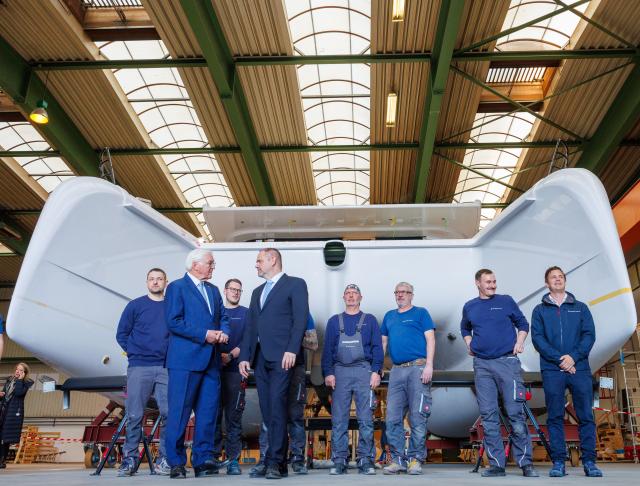 15 April 2026, Mecklenburg-Western Pomerania, Stralsund: German President Frank-Walter Steinmeier (2nd from left) talks to Alexander Badrow (4th from left, CDU), Lord Mayor of the Hanseatic City of Stralsund, during a visit to the Pioneer Yacht company on the Volkswerft shipyard site in front of a catamaran sailing ship under construction, watched by boat builders. As part of his "Local Time Germany" series of visits, the Federal President is moving his official residence to Stralsund until 16.04.2026. Photo: Jens Büttner/dpa