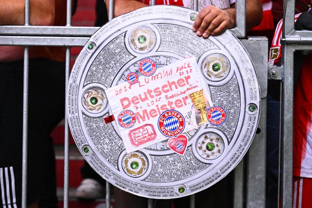19 April 2026, Bavaria, Munich: Fan holds a sign shaped like the German Bundesliga championship trophy ahead of the German Bundesliga soccer match between Bayern Munich and VfB Stuttgart at Allianz Arena. Photo: Tom Weller/dpa - IMPORTANT NOTICE: DFL and DFB regulations prohibit any use of photographs as image sequences and/or quasi-video.