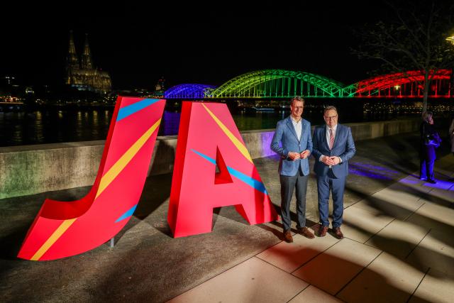 19 April 2026, North Rhine-Westphalia, Cologne: (L-R) Hendrik Wuest, Minister-President of North Rhine-Westphalia, and Torsten Burmester, Mayor of Cologne, stand in front of a large "Yes" sign and the Hohenzollern Bridge illuminated in the Olympic colours. Photo: Christoph Reichwein/dpa