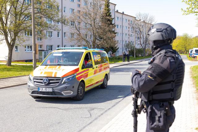 28 April 2026, Saxony, Neustadt: An ambulance drives past a police officer near the scene of the crime after gunshots in the vicinity of an apartment building. Police discovered two dead and one seriously injured person. Photo: Jürgen Lösel/dpa