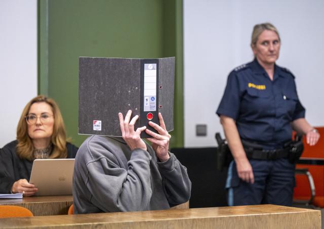 06 May 2026, Bavaria, Munich: The defendant holds a file cover in front of his face at the start of the trial in Munich Regional Court I. The man is on trial for the murder of his stepmother. His lawyer Birgit Schwerdt is sitting on the left. Photo: Peter Kneffel/dpa