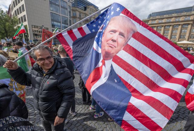 16 May 2026, Hesse, Frankfurt/Main: A man waves a US flag bearing an image of US President Donald Trump during a demonstration at Rathenauplatz in support of the Iranian opposition politician and son of the Shah, Mohammad Reza Pahlavi. Photo: Boris Roessler/dpa