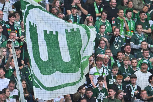 21 May 2026, Lower Saxony, Wolfsburg: VfL Wolfsburg fans are cheering in the stands holding a large flag ahead of the German Bundesliga relegation playoff soccer match between VfL Wolfsburg and SC Paderborn 07 at Volkswagen Arena. Photo: Swen Pförtner/dpa - WICHTIGER HINWEIS: Gemäß den Vorgaben der DFL Deutsche Fußball Liga bzw. des DFB Deutscher Fußball-Bund ist es untersagt, in dem Stadion und/oder vom Spiel angefertigte Fotoaufnahmen in Form von Sequenzbildern und/oder videoähnlichen Fotostrecken zu verwerten bzw. verwerten zu lassen.