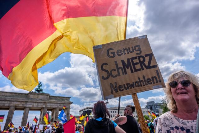 08 June 2026, Berlin: A protester holds a poster with the words "Enough pain, new elections!" in front of the Brandenburg Gate during a demonstration. Organized by the "Project M1llion" movement, the assembly called for the immediate resignation of the current federal government and the implementation of a direct democracy modeled on the Swiss system. Photo: Michael Kappeler/dpa