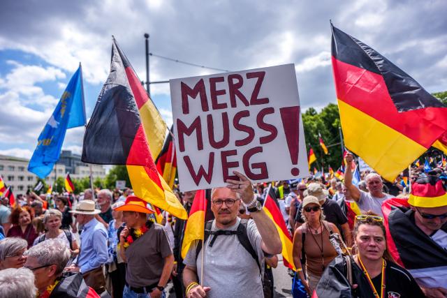 08 June 2026, Berlin: A protester holds a poster with the words "Merz must go!" in front of the Brandenburg Gate during a demonstration. Organized by the "Project M1llion" movement, the assembly called for the immediate resignation of the current federal government and the implementation of a direct democracy modeled on the Swiss system. Photo: Michael Kappeler/dpa
