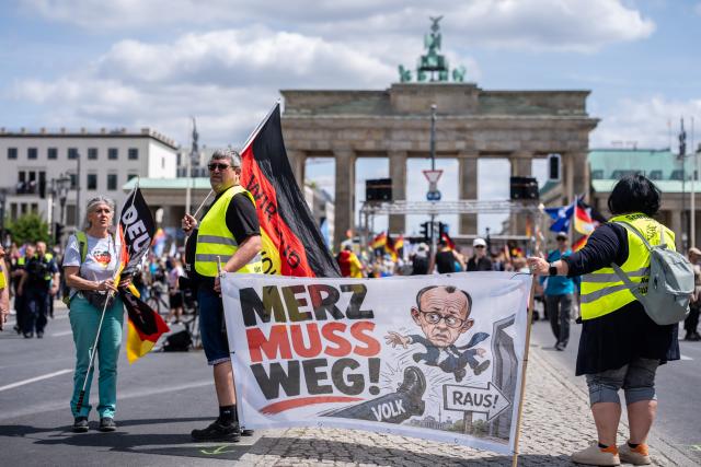 08 June 2026, Berlin: A protester holds a poster with the words  "Merz must go" in front of the Brandenburg Gate during a demonstration. Organized by the "Project M1llion" movement, the assembly called for the immediate resignation of the current federal government and the implementation of a direct democracy modeled on the Swiss system. Photo: Michael Kappeler/dpa