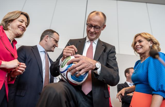 09 June 2026, Berlin: Friedrich Merz (C), Germany's Chancellor, signs an autograph on a soccer ball before the start of the parliamentary group meeting in the German Bundestag. Photo: Michael Kappeler/dpa