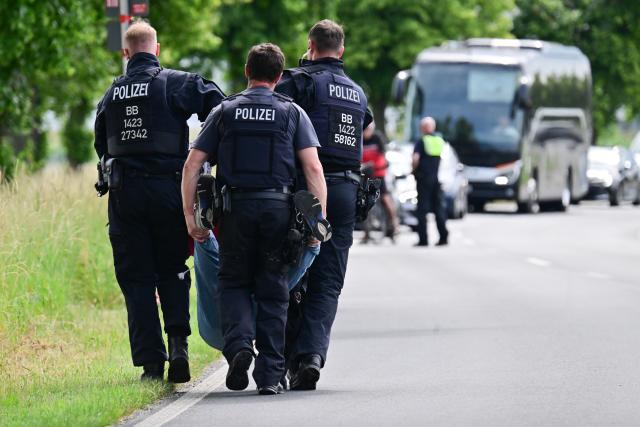 10 June 2026, Berlin, Berlin-Schönefeld: Police officers carry away an activist who had taped herself to an access road during the International Aerospace Exhibition (ILA) at the Berlin ExpoCenter Airport. The ILA begins on June 10 and lasts until June 14. Photo: Sebastian Christoph Gollnow/dpa