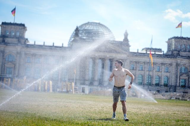 19 June 2026, Berlin: A tourist from Ireland cools off under a lawn sprinkler in front of the Reichstag building amid midsummer temperatures. Photo: Kay Nietfeld/dpa