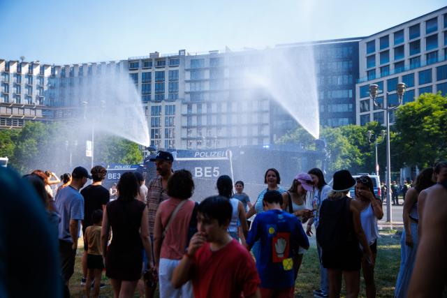 27 June 2026, Berlin: Passersby stand in front of a police water cannon as officers spray water to cool people down during hot weather at Potsdamer Platz in Berlin. Photo: Carsten Koall/dpa