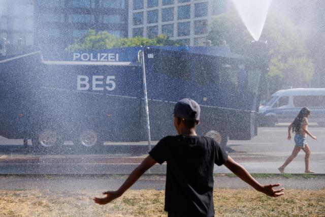 27 June 2026, Berlin: Passersby stand in front of a police water cannon as officers spray water to cool people down during hot weather at Potsdamer Platz in Berlin. Photo: Carsten Koall/dpa