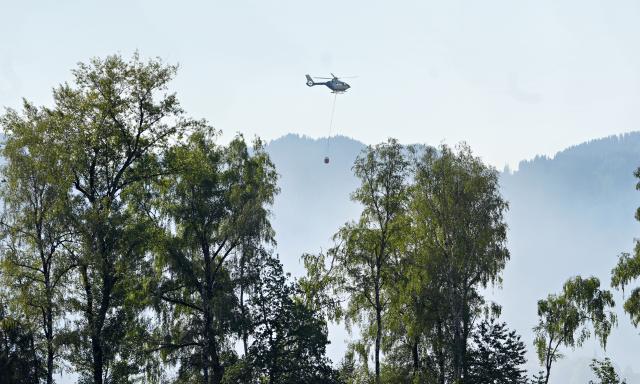 29 June 2026, Bavaria, Übersee: A helicopter carrying water from Lake Chiemsee is flying to a forest and marsh fire while the highway is completely closed. A police spokesperson said the rising smoke is causing reduced visibility. The fire department is also warning the public and asking people to keep their windows and doors closed. Photo: Uwe Lein/dpa