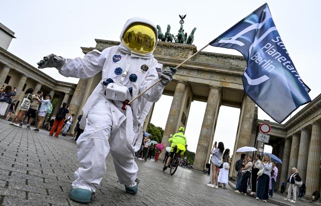 01 July 2026, Berlin: An actress dressed as an astronaut stands in front of the Brandenburg Gate, holding a flag to mark the 10th anniversary of the Planetarium Berlin Foundation. During the anniversary year, a new fulldome production will be shown and special stargazing events will be offered. Photo: Jens Kalaene/dpa